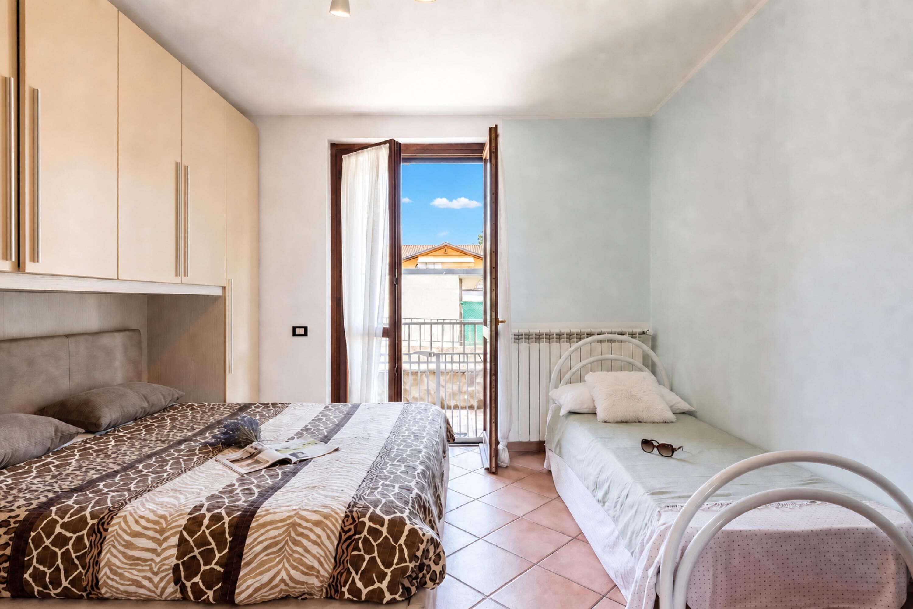 A bedroom with a bed, a smaller bed, a wardrobe, and a window with a view of a building outside.