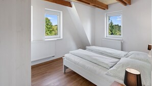 Une chambre avec un plafond en pente, deux fenêtres donnant sur la verdure, un lit avec une literie blanche et une lampe de chevet.
