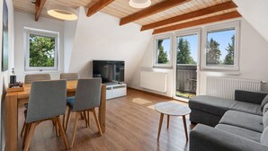 Un appartement moderne de style « loft » comprenant un coin repas, un salon avec un canapé et un téléviseur, et de grandes fenêtres offrant une vue sur les arbres.