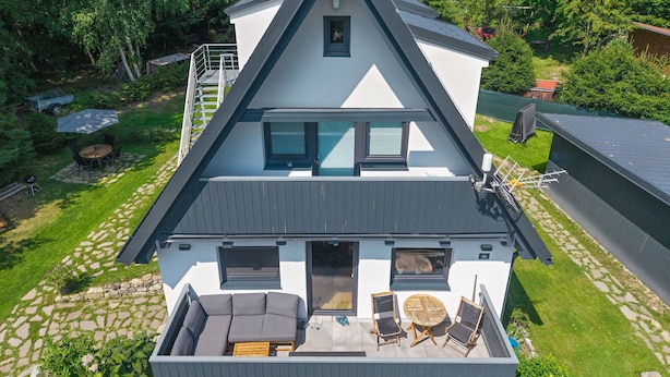 Une maison moderne à deux étages, dotée d’un toit en pente, d’un patio couvert avec des meubles d’extérieur et d’un chemin en pierre menant à l’entrée.