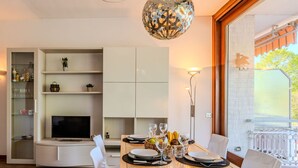 A modern dining area with a wooden table set for four, surrounded by white chairs. A television is mounted on the wall, and there are shelves with various items above the table. - Lovely apartment for 4 guests with WIFI, A/C, TV and terrace (Ispra)