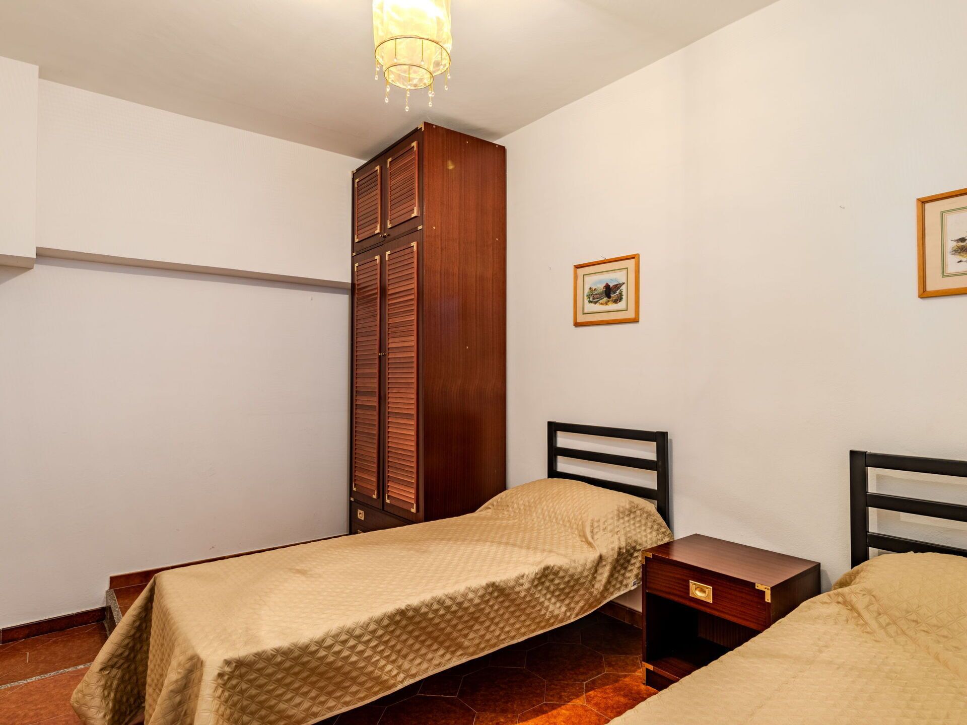 A bedroom with two single beds, a wooden wardrobe, a nightstand, and a hanging light fixture.