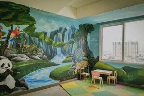 A room with a mural of a forest scene, a window with a city view, and a small table and chair.