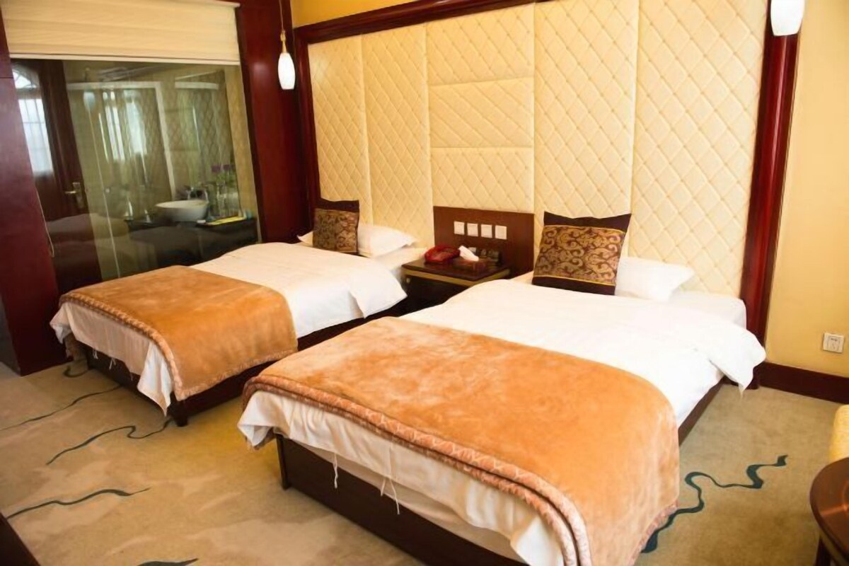 A hotel room with two beds, a large quilted headboard, a bedside table with a lamp, and a view of the bathroom through a glass door.