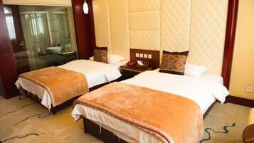 A hotel room with two beds, a large quilted headboard, a bedside table with a lamp, and a view of the bathroom through a glass door.