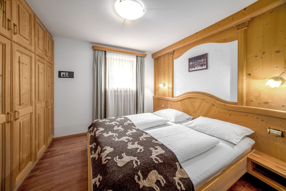 Habitación de hotel con cama de madera, un ventanal con cortinas, armario de madera y lámpara de pared.