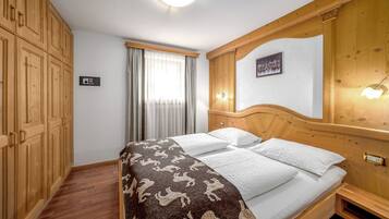 A hotel room with a wooden bed, a large window with curtains, a wooden wardrobe, and a wall-mounted light fixture.