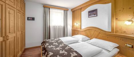 A hotel room with a wooden bed, a large window with curtains, a wooden wardrobe, and a wall-mounted light fixture.