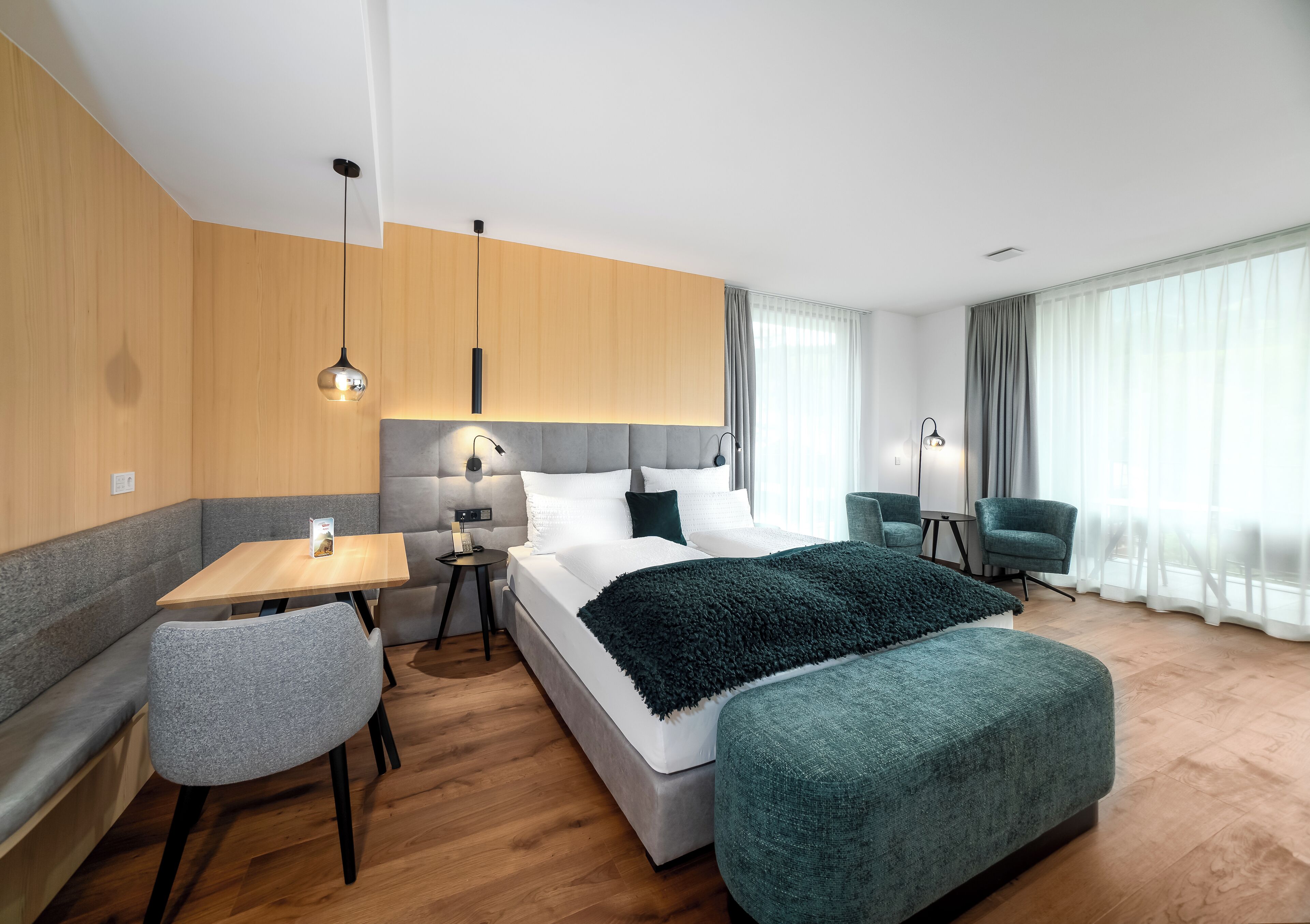 Una habitación de hotel moderna con una cama grande, una zona de estar con mesa y sillas, y vistas a la ciudad a través de amplias ventanas.