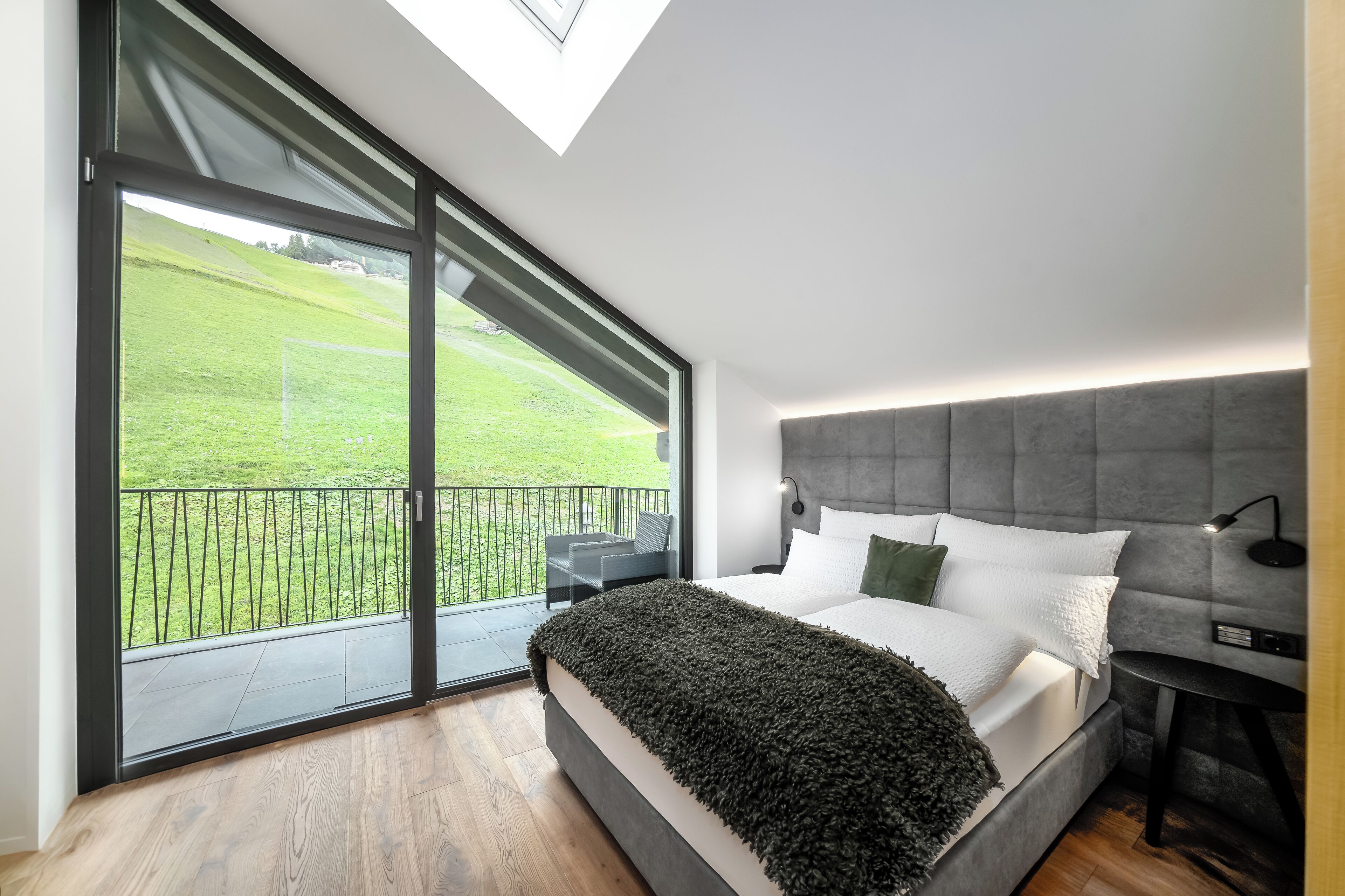 Un dormitorio moderno con una cama grande, un cabecero gris y vistas a un paisaje verde a través de un ventanal corredero.