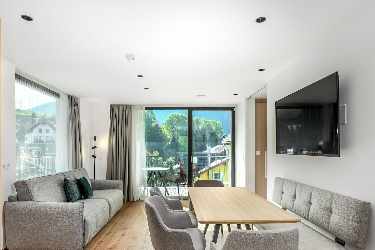 Una sala de estar moderna con un sofá, una mesa de comedor y sillas. Se aprecia un ventanal con vistas a árboles y edificios.