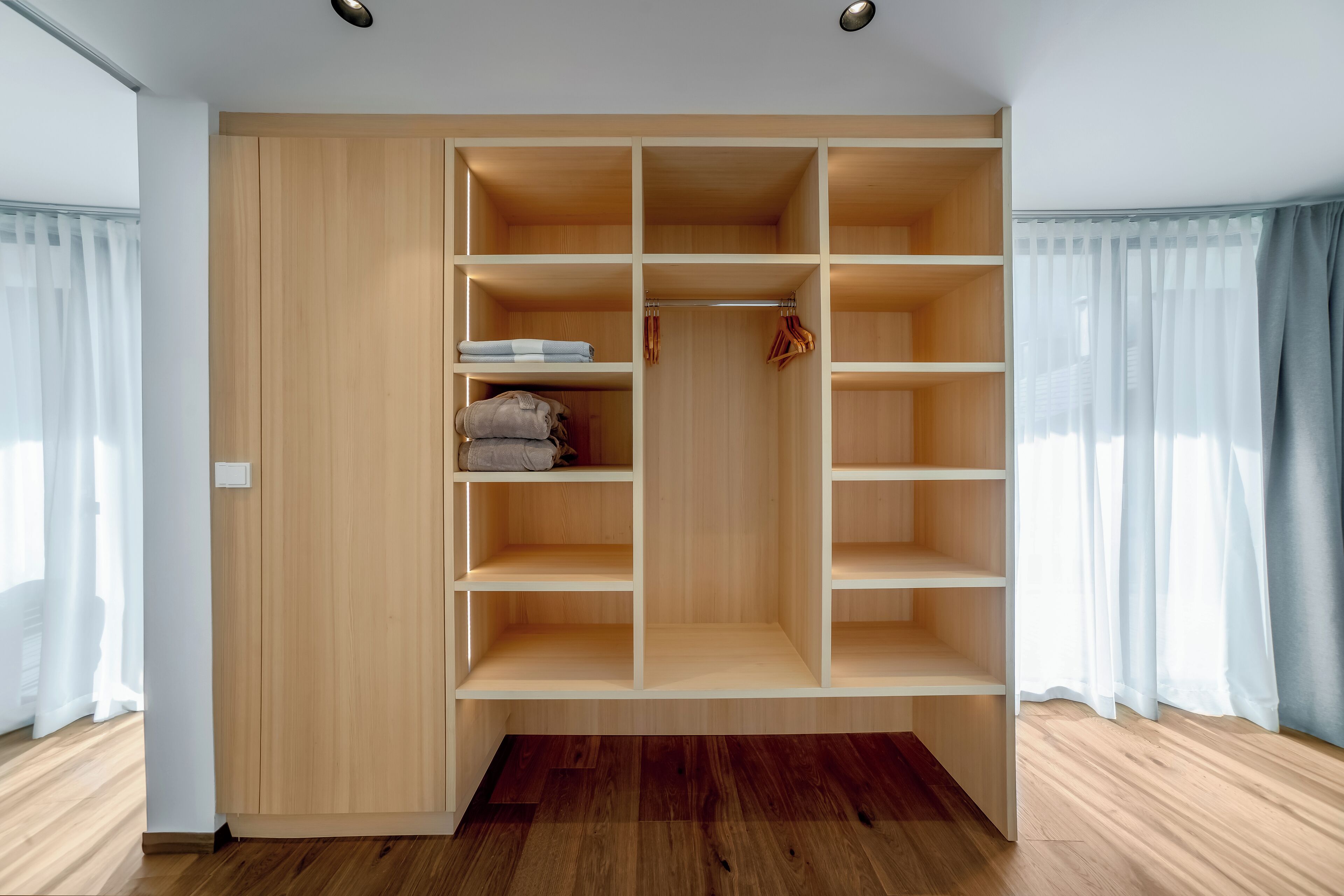 A walk-in closet with multiple shelves and hanging space, wooden flooring, and a window with curtains.