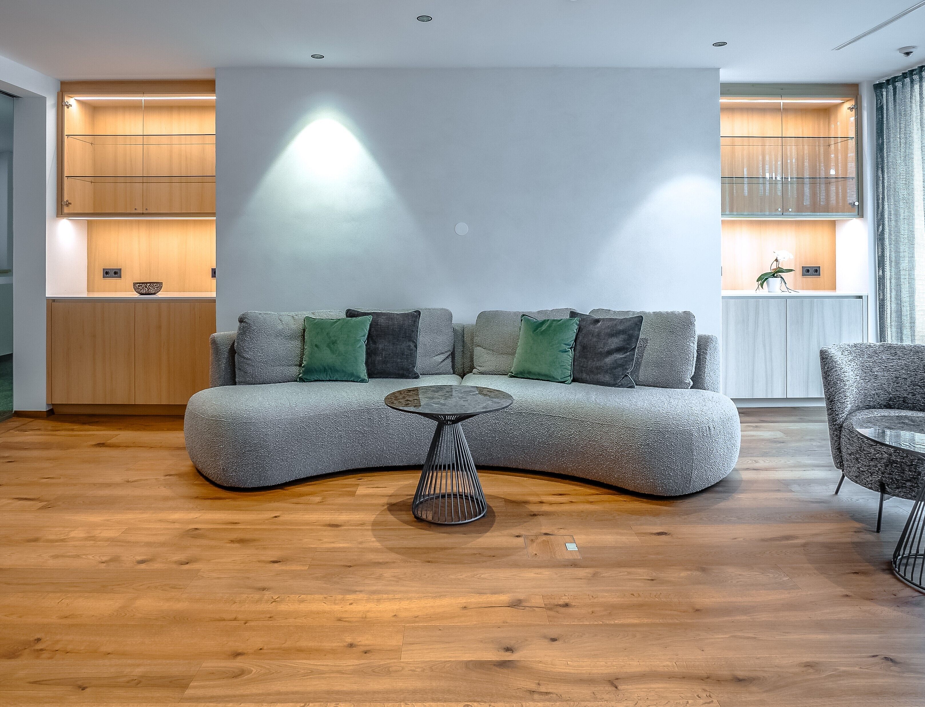 Una sala de estar moderna con un sofá gris curvo, dos cojines verdes y dos grises, una mesa de centro redonda de metal, suelo de madera y armarios empotrados de madera con iluminación integrada.
