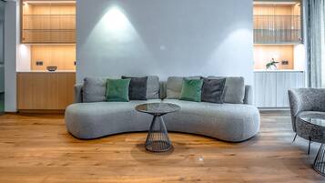 Una sala de estar moderna con un sofá gris curvo, dos cojines verdes y dos grises, una mesa de centro redonda de metal, suelo de madera y armarios empotrados de madera con iluminación integrada.