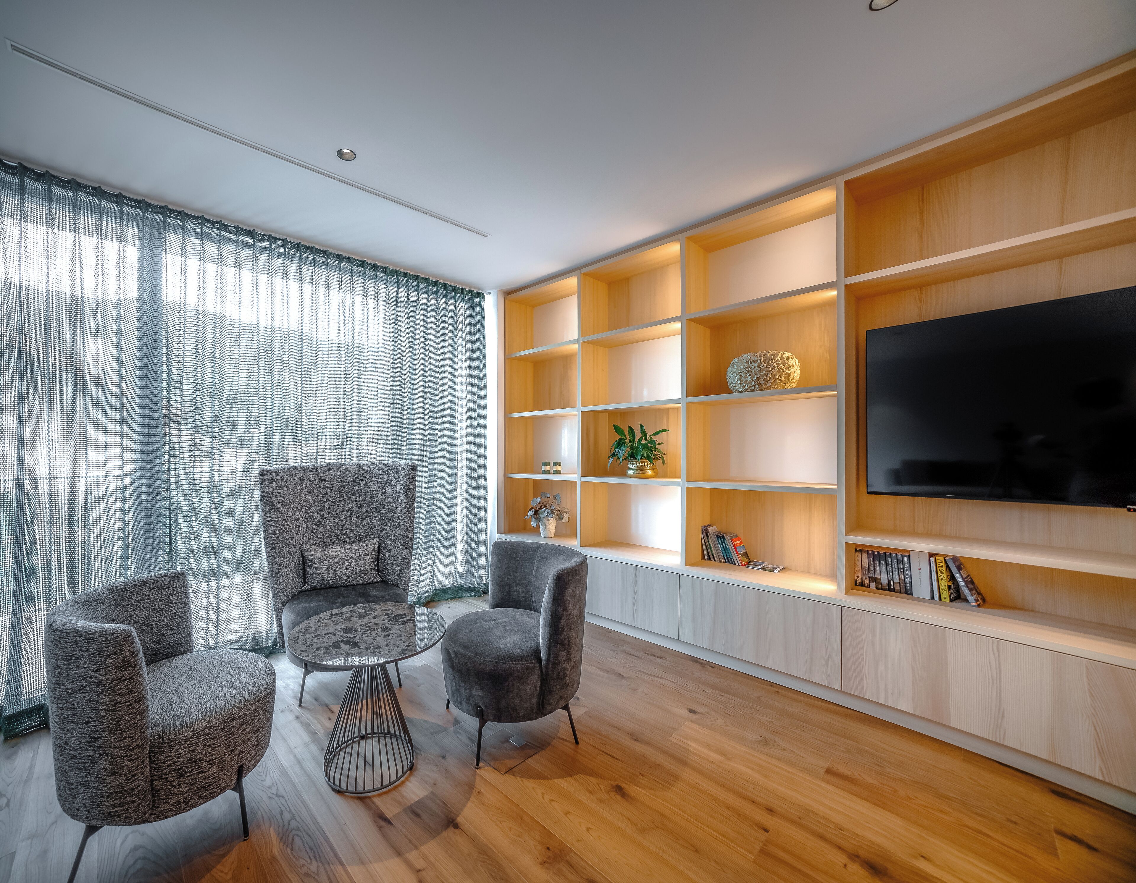 A modern living room with a wooden floor, grey upholstered chairs, a round coffee table, and a built-in wooden shelving unit with a flat-screen TV.