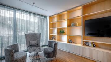 A modern living room with a wooden floor, grey upholstered chairs, a round coffee table, and a built-in wooden shelving unit with a flat-screen TV.