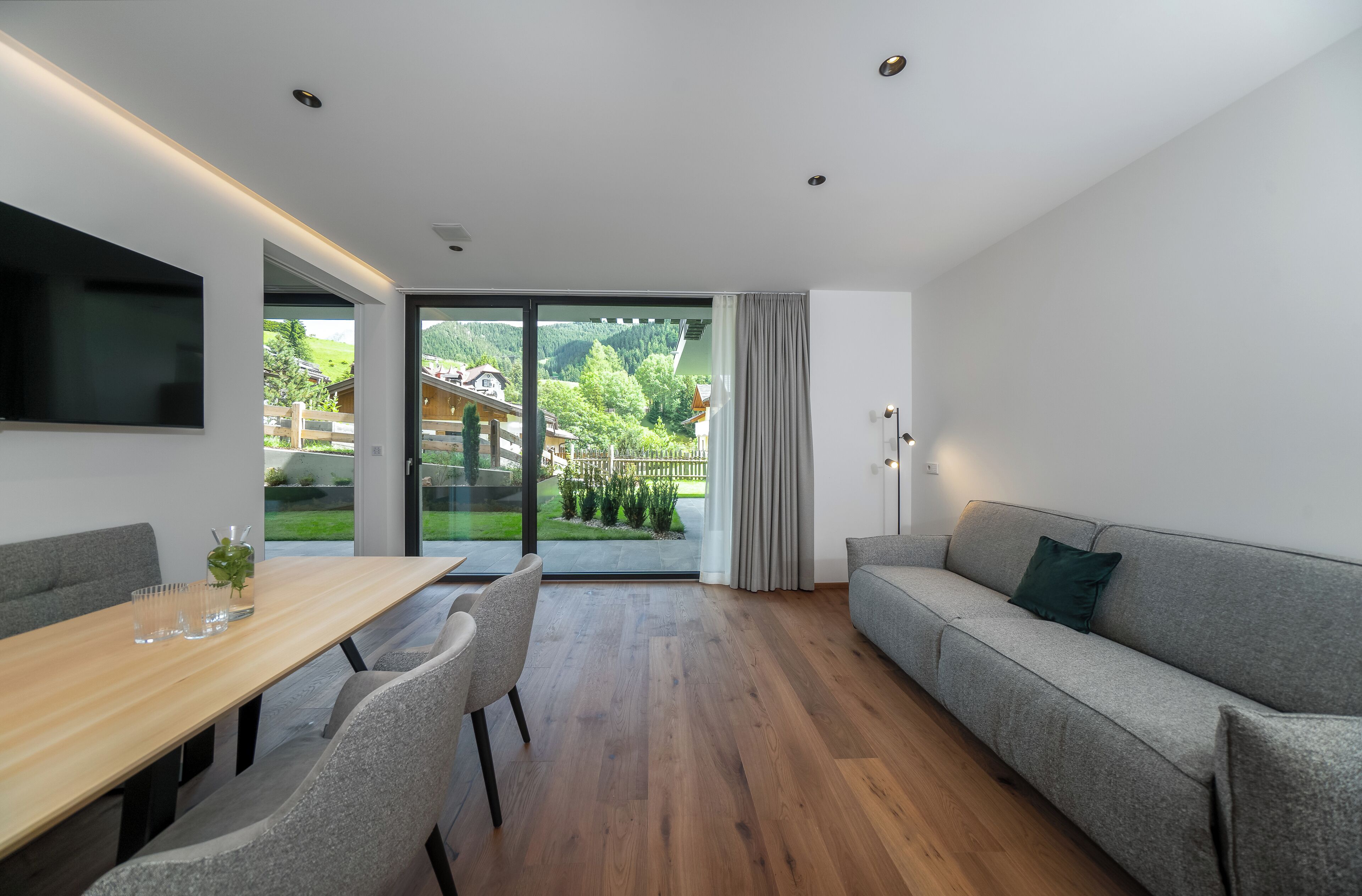 A modern living room with a wooden dining table, grey sofa, and a TV mounted on the wall.