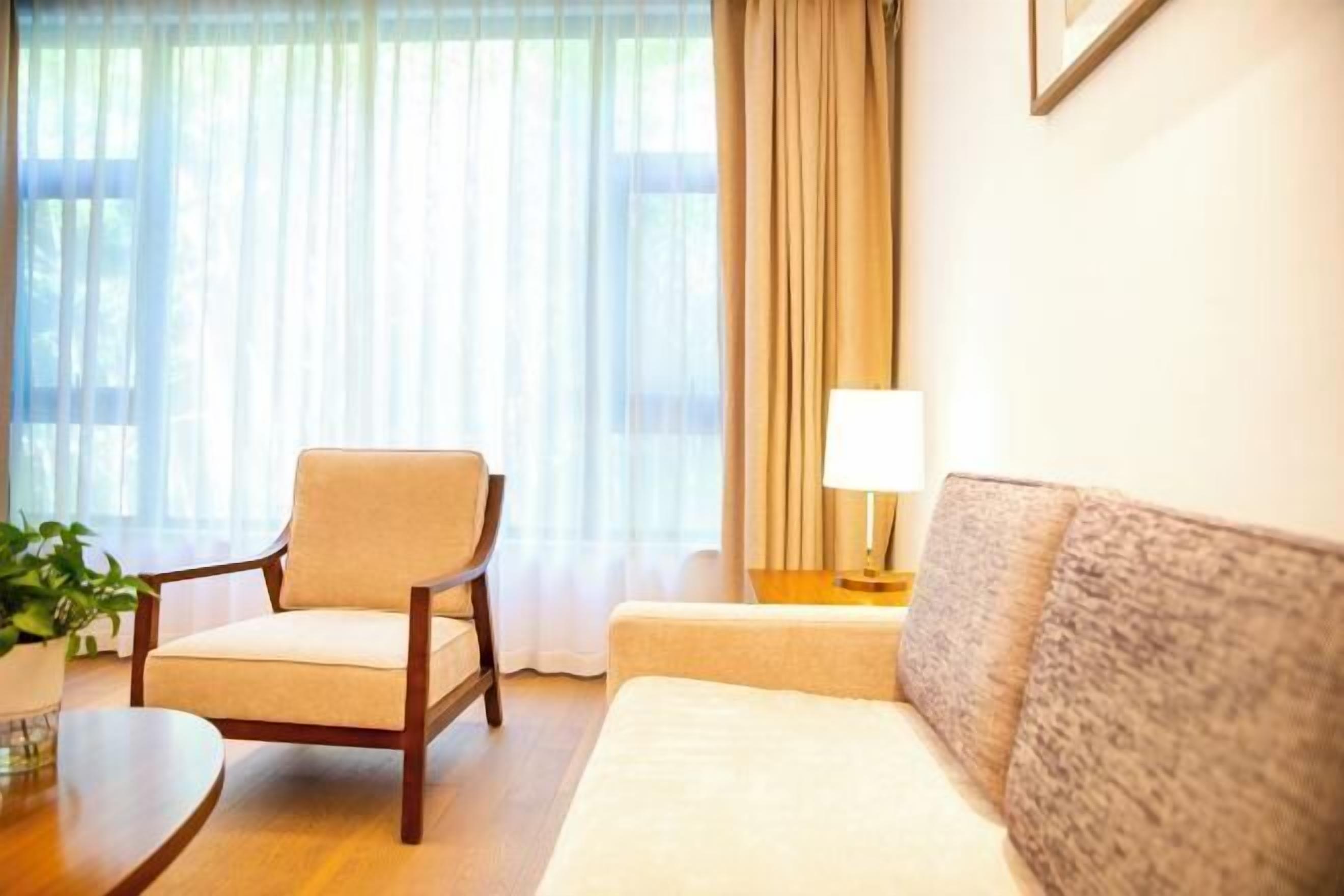 Ein Hotelzimmer mit Sofa, Sessel, Nachttisch, Lampe und einem Fenster mit Vorhängen.