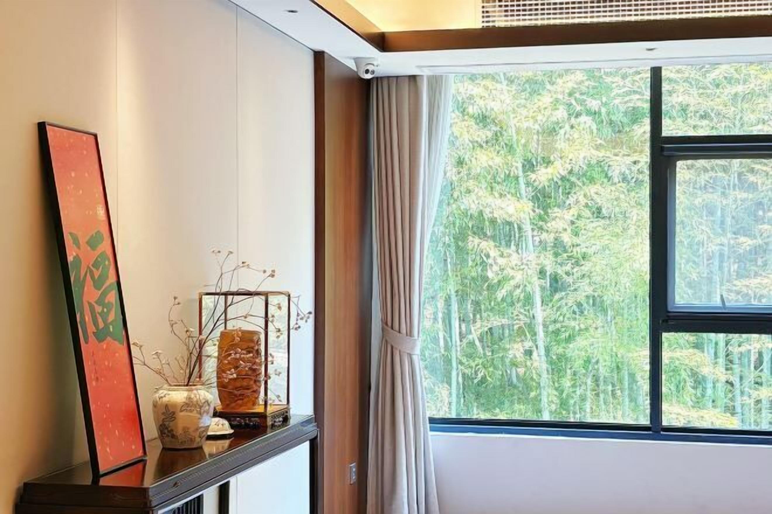 A room with a large window, a wooden shelf with decorative items, and a view of bamboo trees outside.