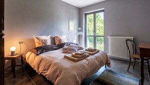 A bedroom with a large bed, a desk, a chair, a radiator, and a window with a view of greenery.