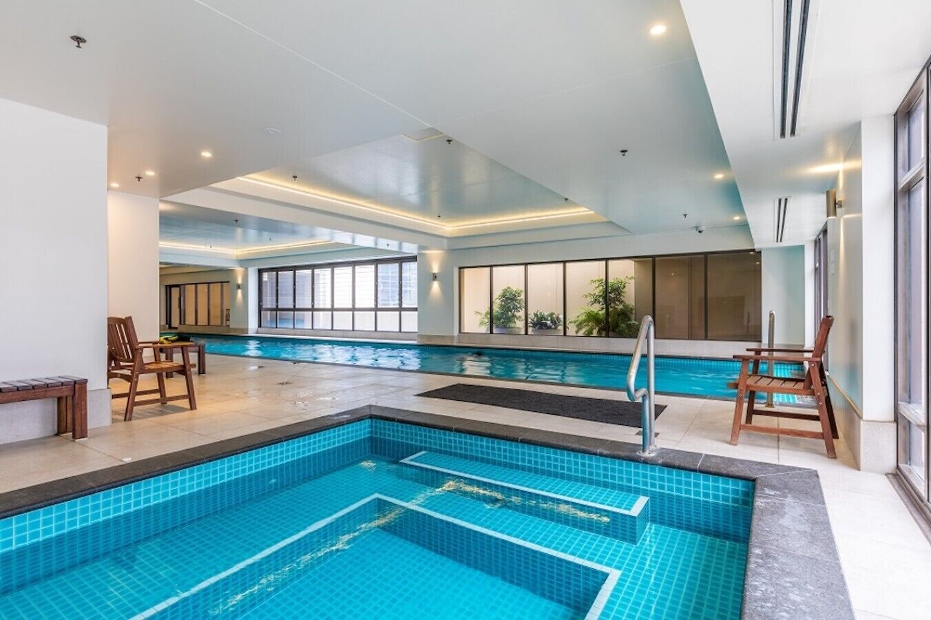 Une grande piscine intérieure à l’eau d’un bleu limpide, entourée de chaises et de tables en bois, et dotée de grandes fenêtres qui laissent entrer la lumière naturelle.