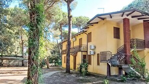 A yellow building with a balcony and air conditioning units, surrounded by trees and a concrete path. - Lovely apartment for 4 people with A/C, TV and pets allowed (Rosolina)