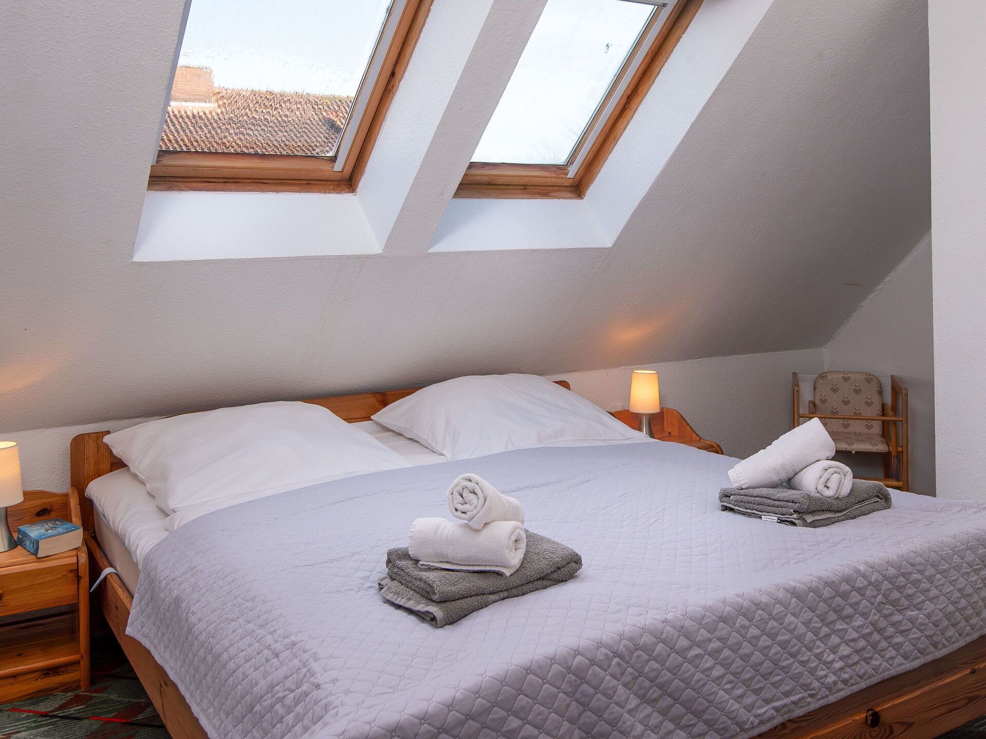 A bedroom with a sloped ceiling, two skylights, a bed with white linens, a bedside table with a lamp, and a chair with a heart pattern.