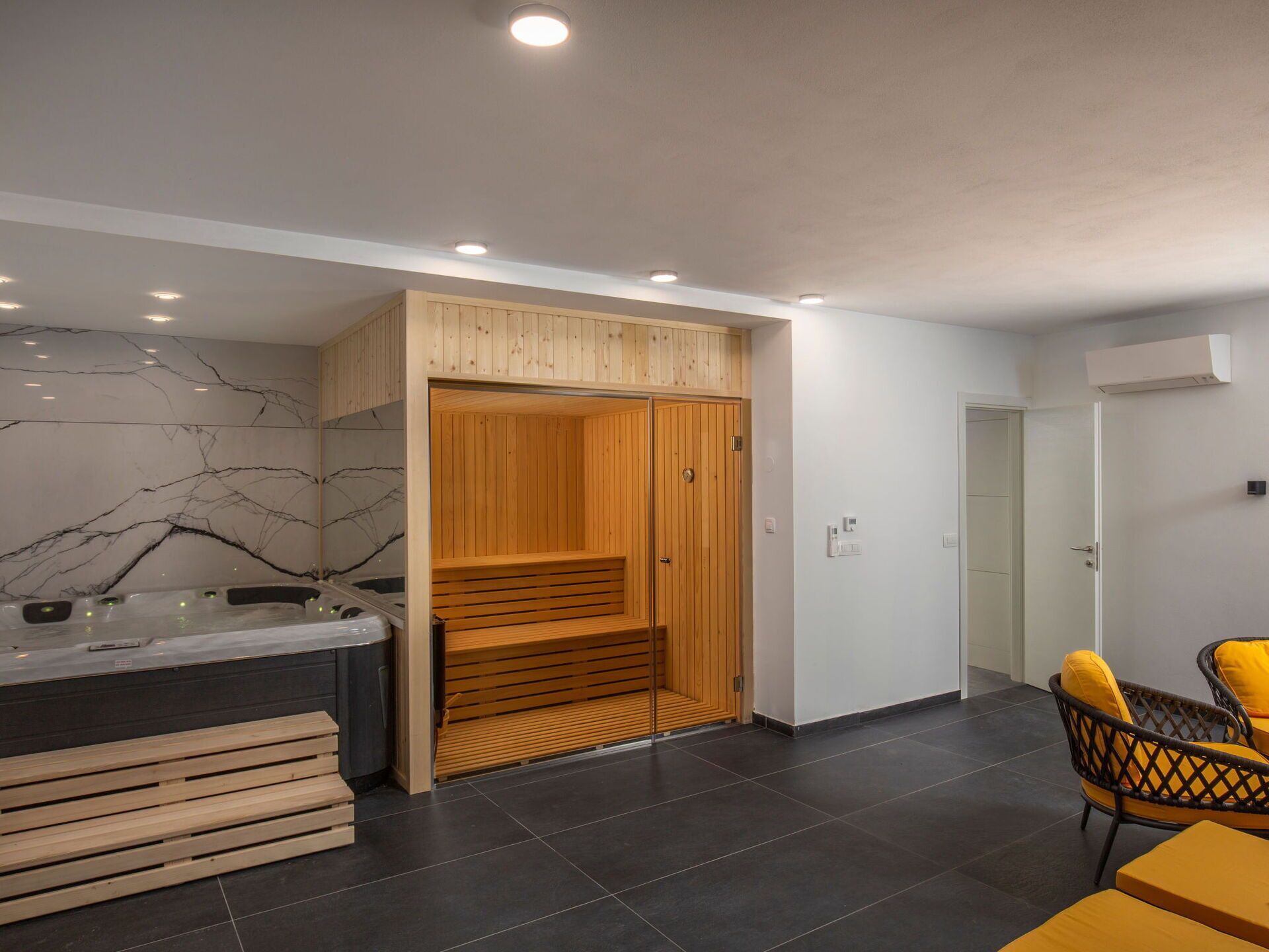Une pièce moderne équipée d’un sauna en bois, d’une table de massage et d’un coin salon avec des coussins jaunes.