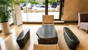 Un vestíbulo con un banco de piedra, sillas de mimbre, una planta en maceta y vistas de coches estacionados a través del vidrio.