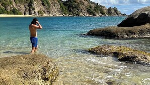 Une personne se tient sur une côte rocheuse, au bord d’une mer bleue et limpide, avec un paysage vallonné en arrière-plan.