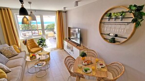 Una sala de estar con sofá, sillón, mesa de comedor, televisor y un balcón con vistas al mar.