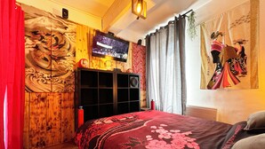Een slaapkamer met een houten kledingkast, een aan de muur bevestigde televisie en een bed met een rood en zwart bloemenbeddengoed.