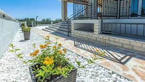 A modern building with stone and concrete exterior, a balcony with metal railings, and a pathway with white pebbles and yellow flowers. - Stunning private villa for 8 guests with A/C, hot tub, WIFI, private pool, TV and terrace (Zadar)
