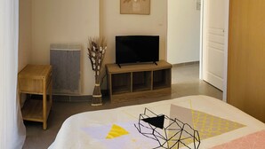 A bedroom with a bed, a television on a wooden stand, a radiator, a vase with dried plants, and a painting on the wall. - Maison Moderne Avec Jardin (Saint Xandre)