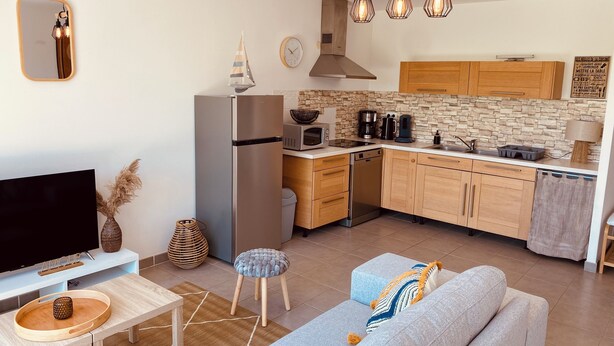 A modern kitchen with wooden cabinets, a stainless steel refrigerator, and a built-in oven. There is a dining area with a wooden table and a sofa. - Maison Moderne Avec Jardin (Saint Xandre)