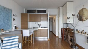 A modern kitchen with wooden cabinets, a white countertop, and a central island. There is a dining area with striped chairs and a table.