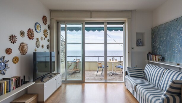 A living room with a TV, bookshelves, a striped sofa, and a view of the sea through large glass doors.