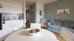 Une cuisine moderne avec des meubles blancs, une table à manger sur laquelle se trouve un saladier rempli de nourriture, un canapé gris et un cactus accroché au mur.