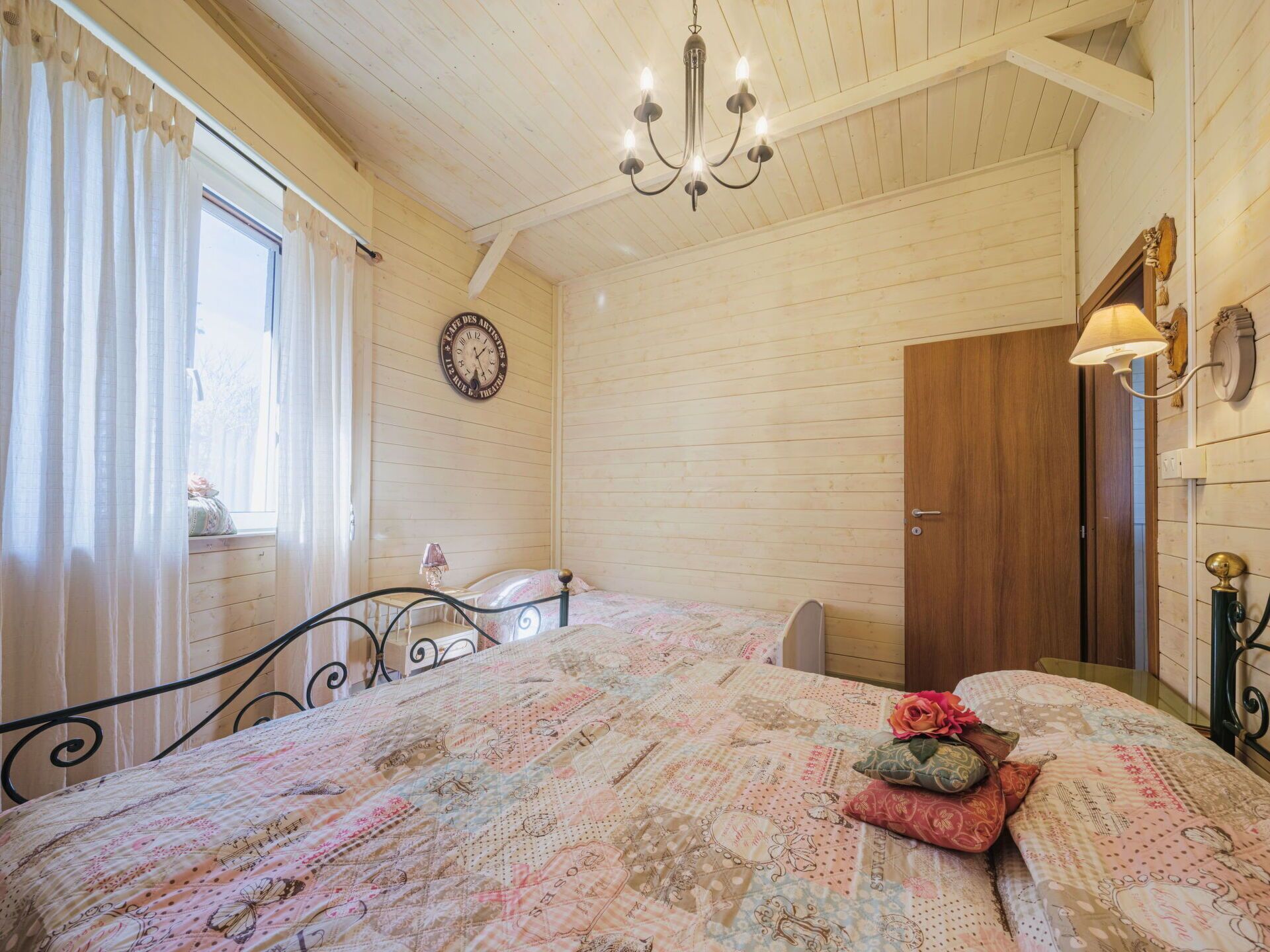 A cozy bedroom with a bed, a wooden door, a chandelier, and a clock on the wall.