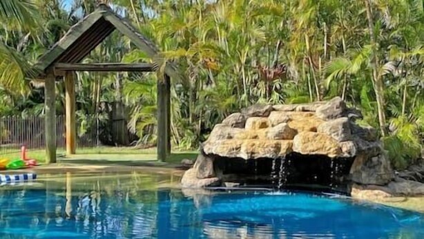 Une piscine agrémentée d’une cascade artificielle en roche, entourée d’une végétation luxuriante et d’une pergola en bois.