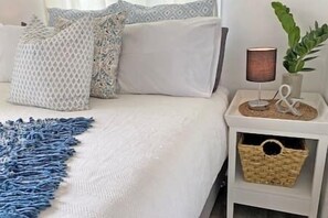 A neatly made bed with a white duvet, a blue fringe throw, and patterned pillows. There is a bedside lamp and a small plant on a nightstand. - Belles on Ballow - Possum Lodge (Amity Point)