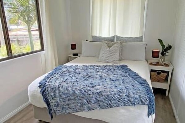 A bedroom with a bed, a nightstand, a lamp, and a window with curtains. - Belles on Ballow - Possum Lodge (Amity Point)
