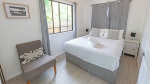 Un dormitorio ordenado con cama, una silla, mesita de noche, lámpara y ventana con cortinas.