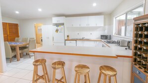 Una cocina moderna con armarios blancos, una isla central con taburetes, una zona de comedor con mesa de madera y una estantería para vinos.