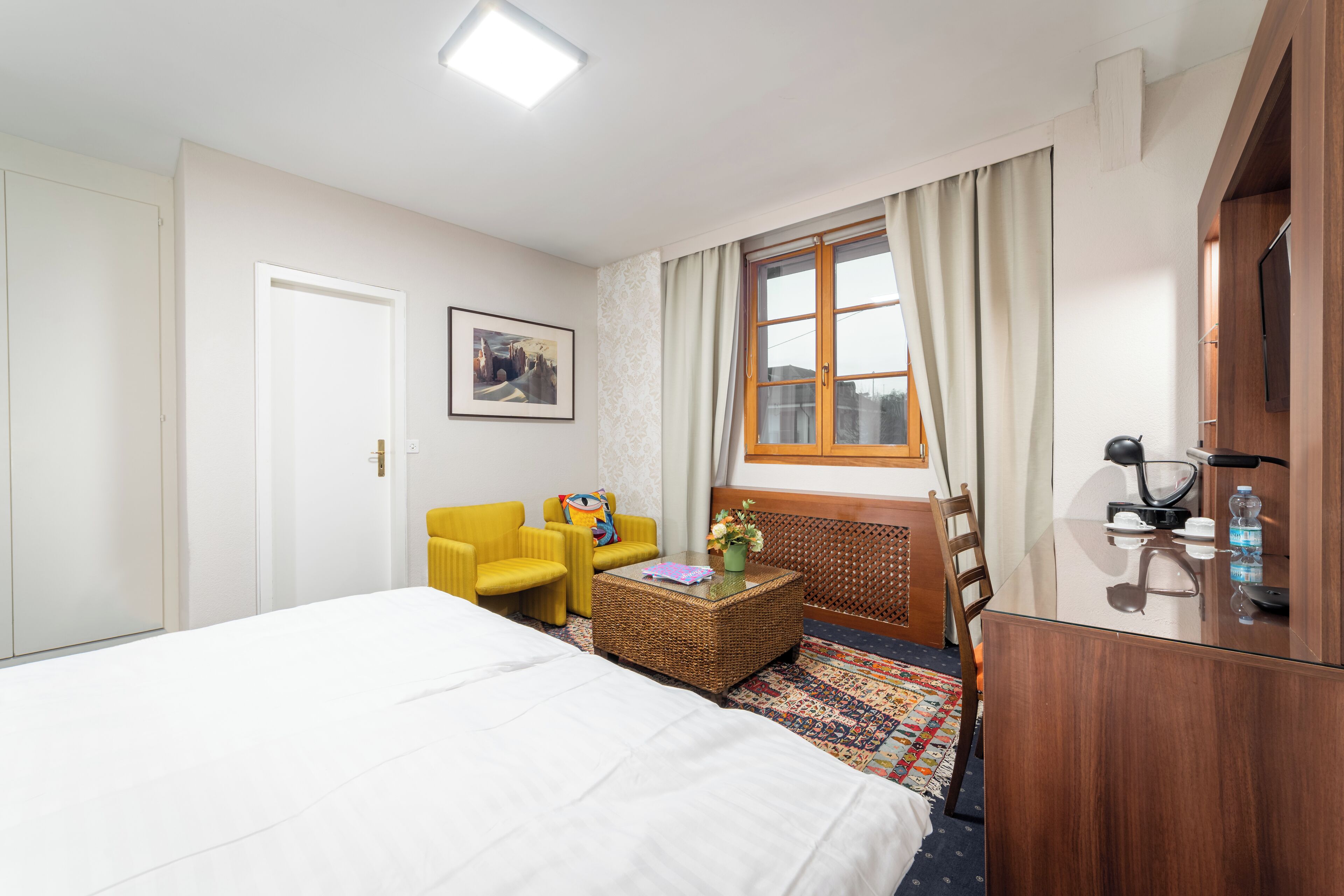 A hotel room with a bed, a yellow armchair, a wicker coffee table, a wooden desk, and a window with curtains.
