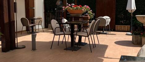 Een terras buiten met een tafel en stoelen, een grote parasol en een decoratieve plantenbak met bloemen.