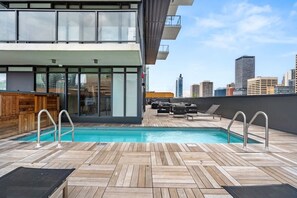Una piscina en la azotea con cubierta de madera, sillas de descanso y el horizonte de la ciudad al fondo.