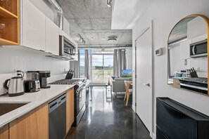 Una cocina moderna con estufa, cafetera y hervidor, un comedor con mesa y sillas, y un amplio ventanal con vistas a la ciudad.