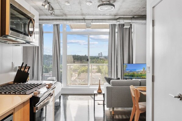 Una cocina moderna con estufa, horno y microondas, un sofá gris, un televisor y un balcón con vista a la ciudad.