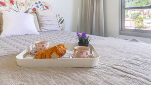Un lit bien fait, avec une tête de lit à motifs floraux, un plateau avec des croissants et des tasses de café, et une vue sur une colline verdoyante par la fenêtre.